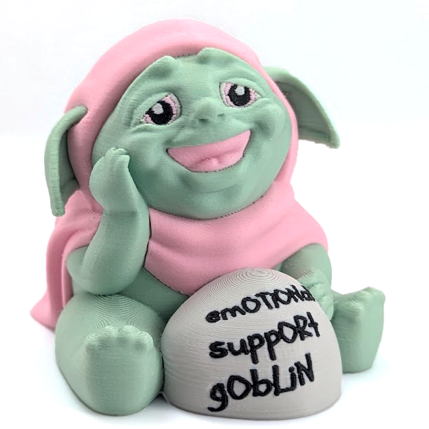 An adorable goblin in a pink cloak, with a wide toothless smile and kind eyes, sits next to a rock that says "emotional support goblin".