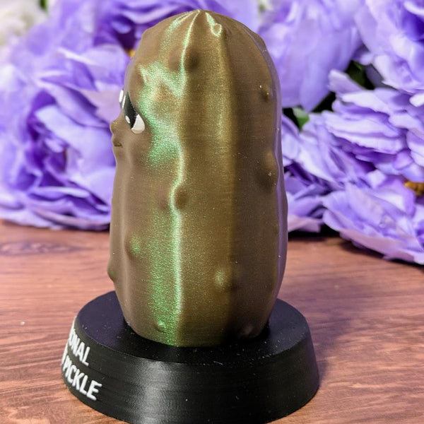 Side view of Emotional Support Pickle figurine with glossy green surface and subtle sparkle, in front of purple flowers.