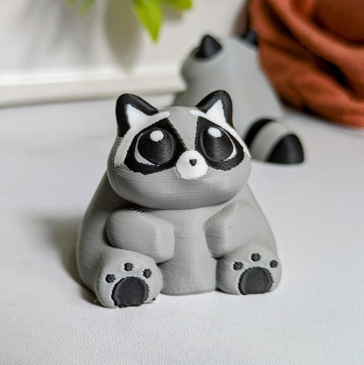 A plump raccoon figurine with soft round features and tiny paws resting contentedly on his belly.