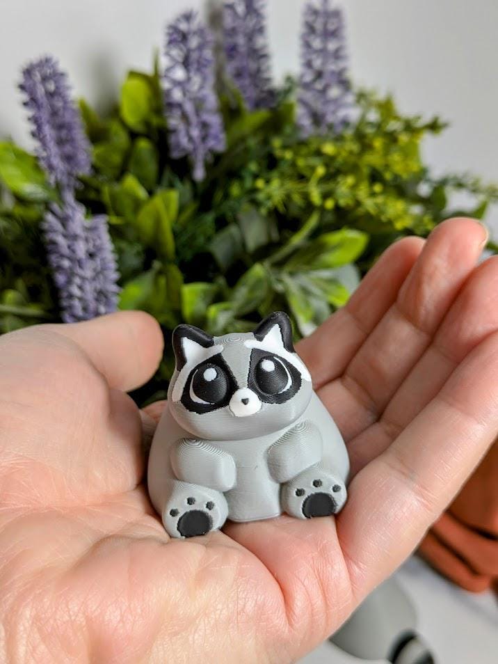 A hand holding the fat raccoon figurine, emphasizing his squishy shape and peaceful little face.