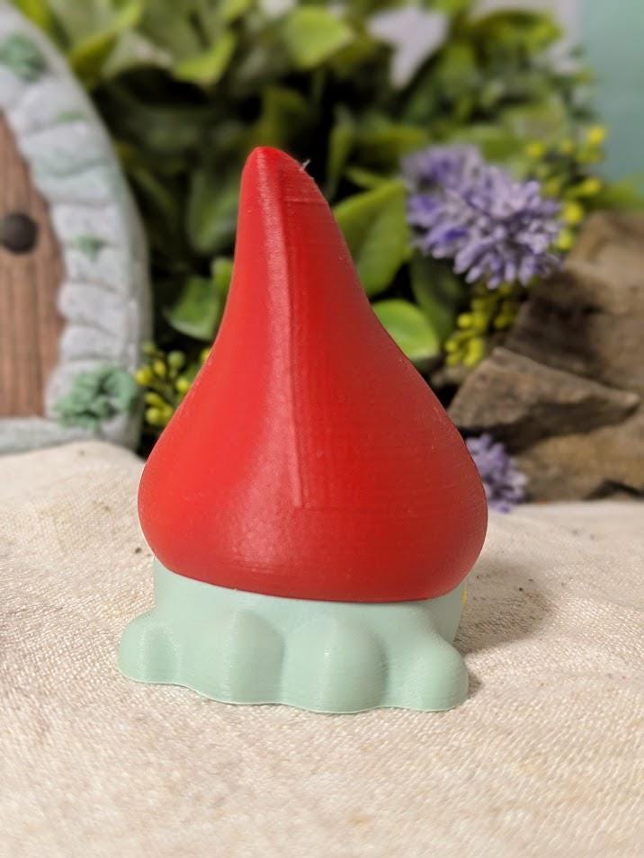 Back view of frog gnome goblin figurine with tall red hat and pale green base, posed on neutral fabric.