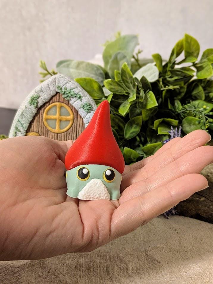 Frog gnome goblin in a large red hat, sitting in a hand to show scale.