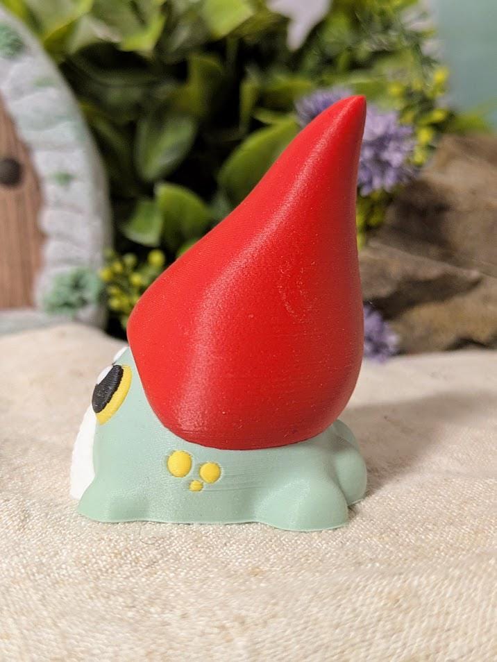 Angled side view of frog gnome goblin figurine showing the curve of its tall red hat and rounded green body.