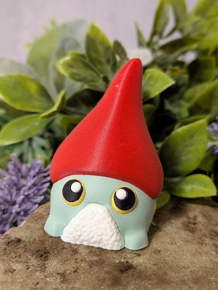 Green gnome frog goblin figurine in tall red hat, posed in front of greenery and florals on a stone surface.