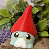 Green gnome frog goblin figurine in tall red hat, posed in front of greenery and florals on a stone surface.