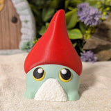 Front view of wide-eyed gnome frog goblin with dramatic red hat, standing on beige cloth with soft focus background.