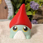 Front view of wide-eyed gnome frog goblin with dramatic red hat, standing on beige cloth with soft focus background.