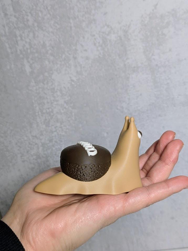 Hand holding a cupcake snail figurine showing its chocolate frosting shell and signature white swirl, resembling a Hostess cupcake.