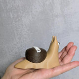 Hand holding a cupcake snail figurine showing its chocolate frosting shell and signature white swirl, resembling a Hostess cupcake.