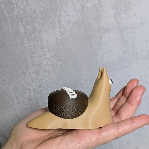 Hand holding a cupcake snail figurine showing its chocolate frosting shell and signature white swirl, resembling a Hostess cupcake.