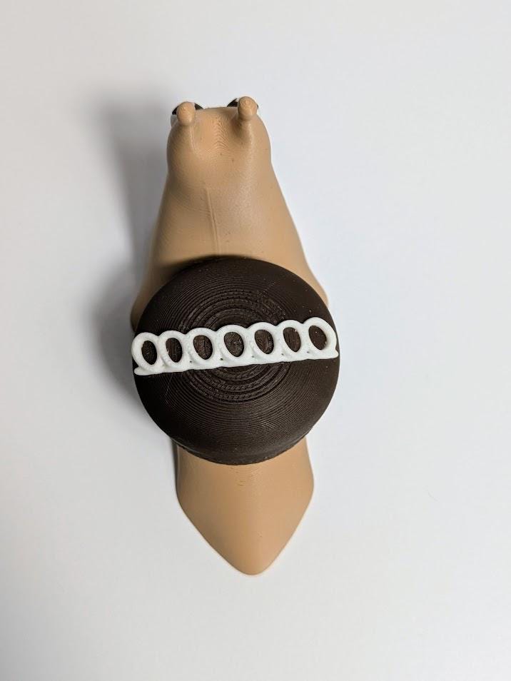 Overhead view of a Hostess-inspired cupcake snail with a tan body and chocolate-frosted shell featuring a white icing swirl.