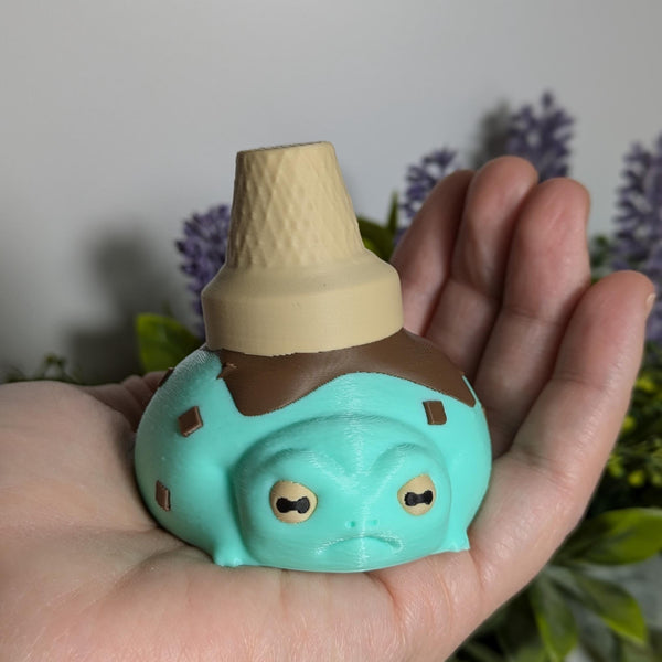 The ice cream frog resting in a hand, looking like it just got scooped from a fairy garden sundae bar.