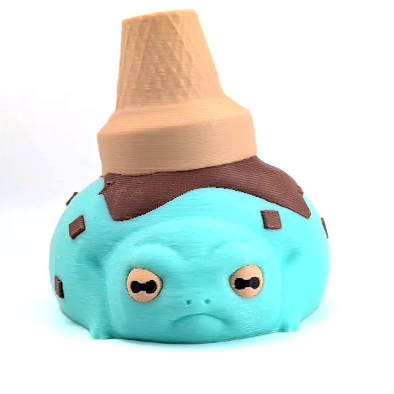 A scoop of mint chocolate chip ice cream that is actually a frog! This grumpy frog has an ice cream cone perched on his back, along with chocolate syrup.
