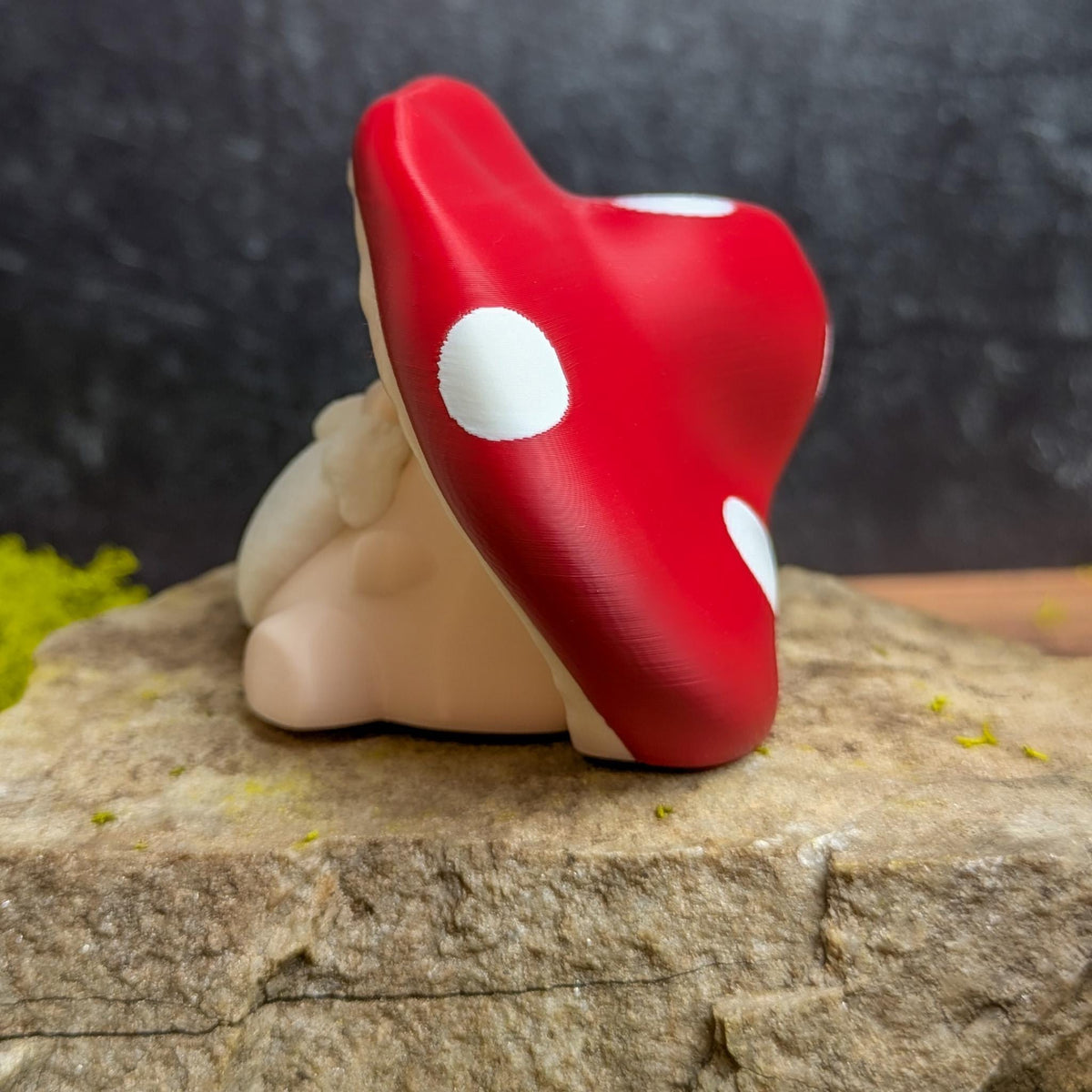 Left side profile of the mushroom man figurine, highlighting the curve of the red mushroom cap and soft body details, placed on textured rock.