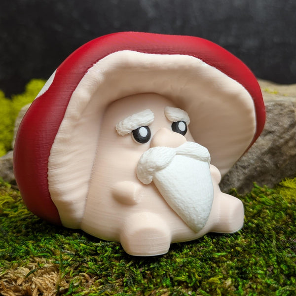 Angled view of a whimsical old mushroom man figurine with a red cap, thick white eyebrows, and a flowing white beard, sitting on moss next to a rock.