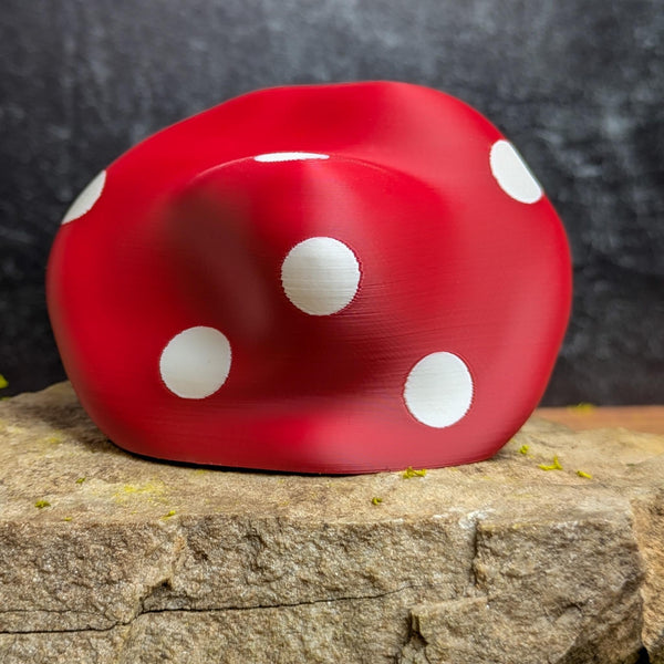 Rear view of the old mushroom man figurine, showing the red mushroom cap with bold white polka dots, resting on a rock slab.
