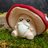 Front view of the mushroom man figurine with an expressive face, white mustache and eyebrows, seated against a mossy background.
