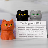 Three cat figurines with a humorous description card labeled 'The Judgmental Cat' on a blurred background.