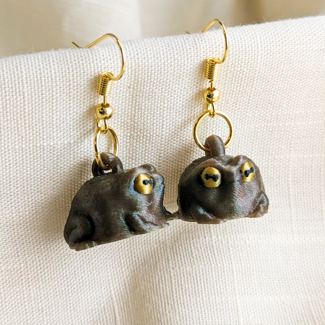 Pair of judgmental frog earrings resting on a cream linen cloth with gold hooks and olive shimmer finish