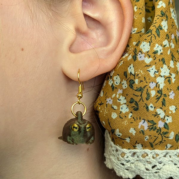 Judgmental frog earring dangling from an ear, showing size and gold hook attachment