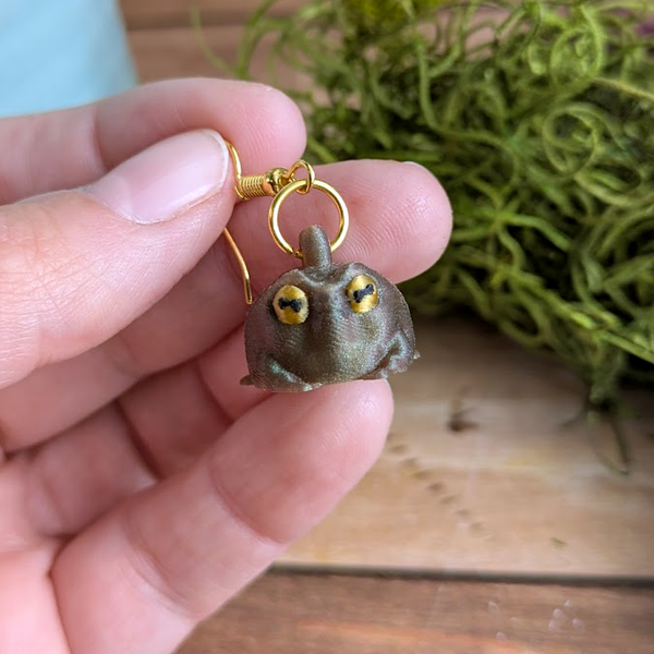 Judgmental frog earrings held in hand with hooks visible, showing shimmer and facial detail