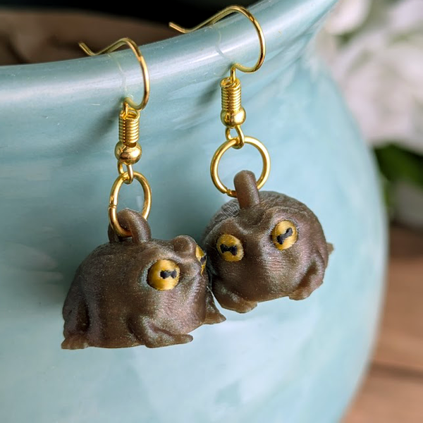 Judgmental frog earrings hanging on a textured vase surrounded by moss, styled in a cottagecore display