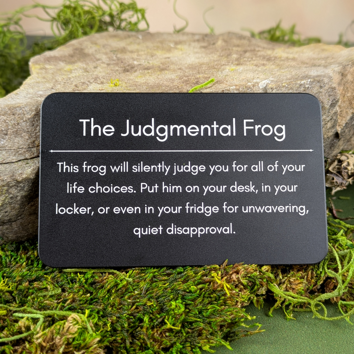 Black metal card with text 'The Judgmental Frog' and a description on a mossy background