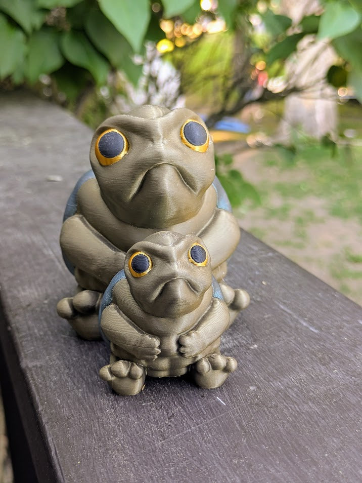 Front-facing view of both large and small Judgmental Turtle™ figurines together on a wood surface.