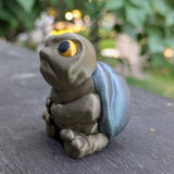 Three-quarter angle of New Sculpt Judgmental Turtle™ with arms resting on legs, seated on wooden surface.