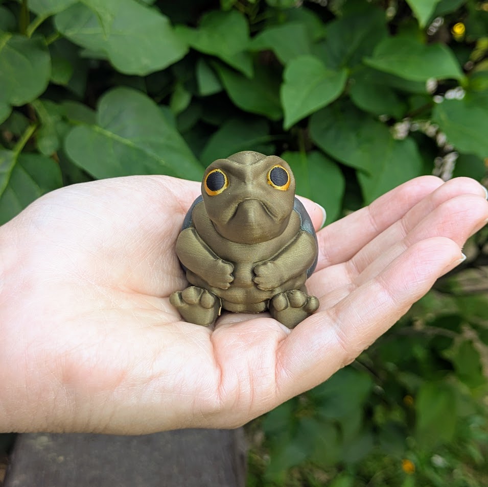 Small Judgmental Turtle™ held in hand for size reference with leafy background.