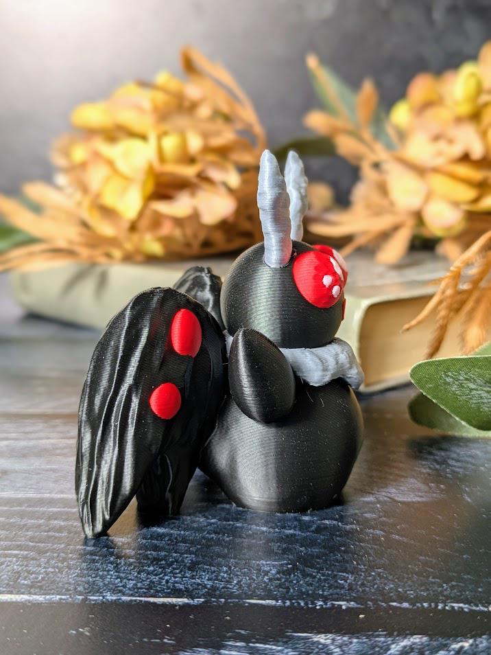 Angled view of Mothman figurine with red eyes and black wings, posed on dark wood with autumn leaves in the background.