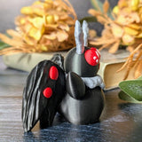 Angled view of Mothman figurine with red eyes and black wings, posed on dark wood with autumn leaves in the background.