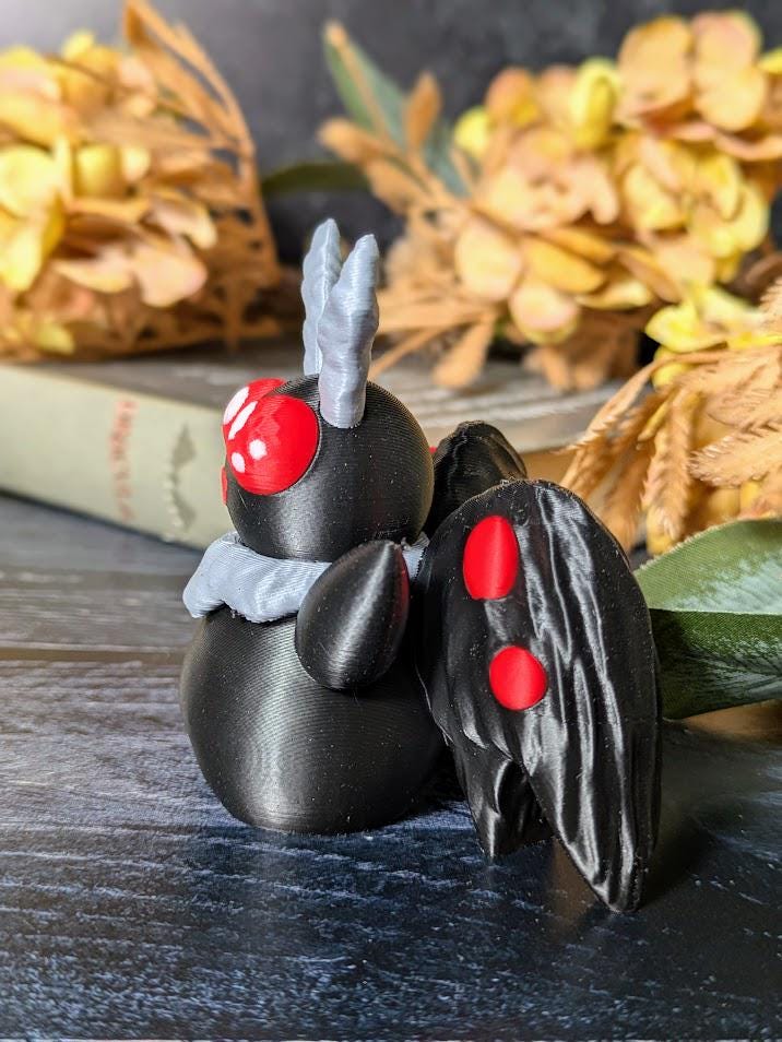 Side view of Mothman with glowing red eyes and folded wings, seated on tabletop with harvest-style decor.