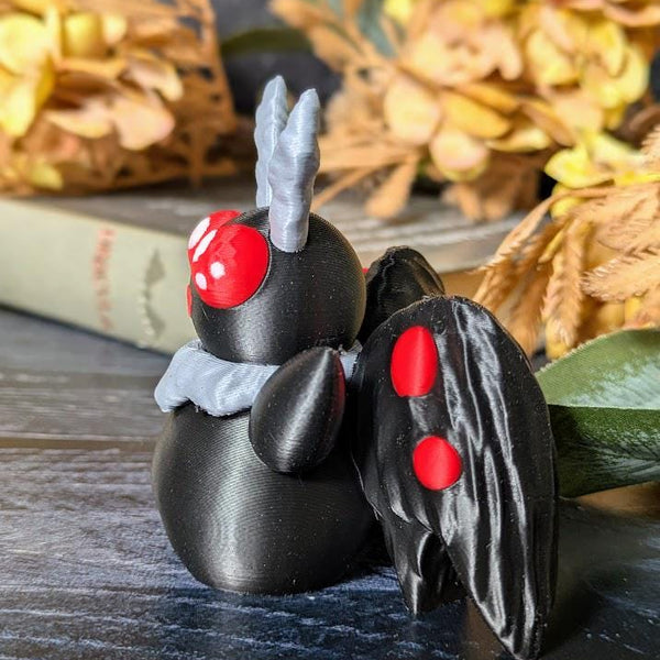 Side view of Mothman with glowing red eyes and folded wings, seated on tabletop with harvest-style decor.