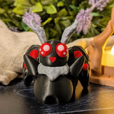 Mothman figurine on dark wood surface with lavender and warm light in background, creating a nighttime cottagecore vibe.