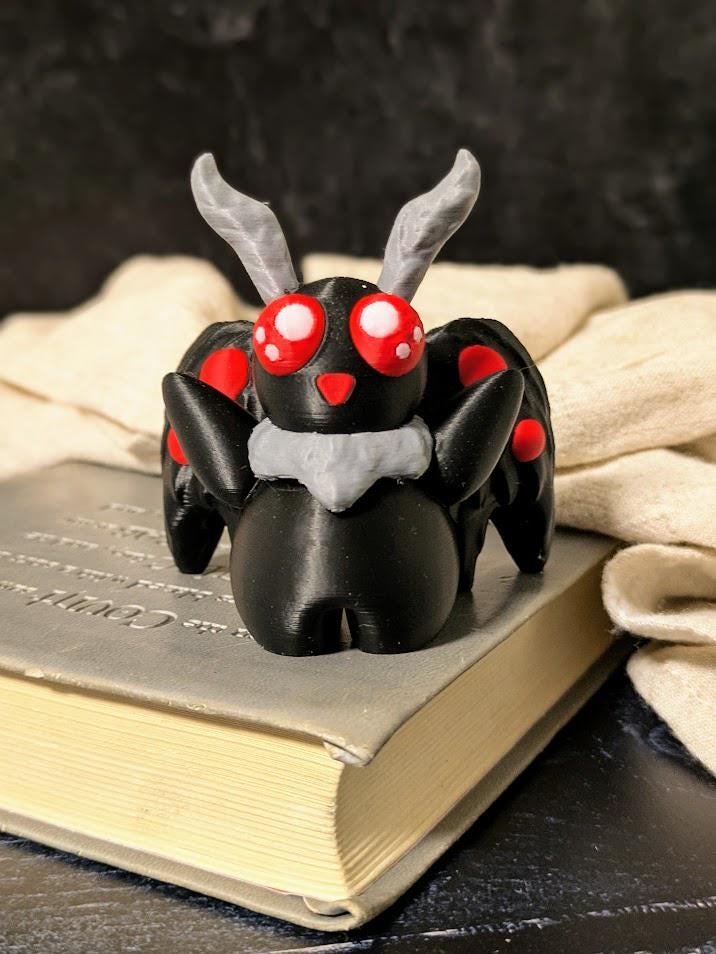 Mothman figurine resting on a book with cozy fabric and candlelit backdrop, red eyes glowing.