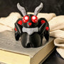 Mothman figurine resting on a book with cozy fabric and candlelit backdrop, red eyes glowing.