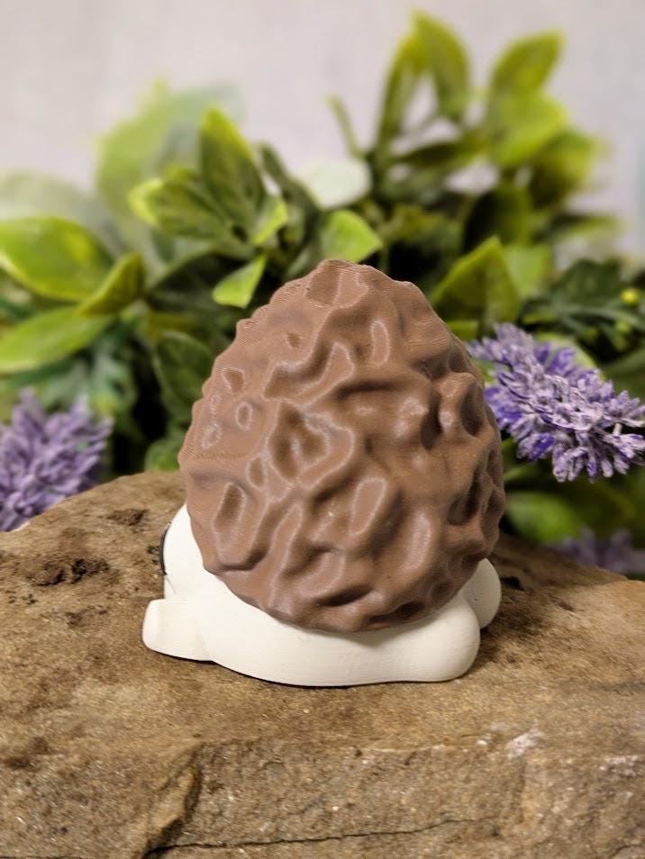 Side view of mushroom figurine with morel-style cap and white base, posed on stone with leafy floral backdrop.