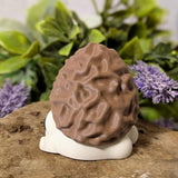 Side view of mushroom figurine with morel-style cap and white base, posed on stone with leafy floral backdrop.