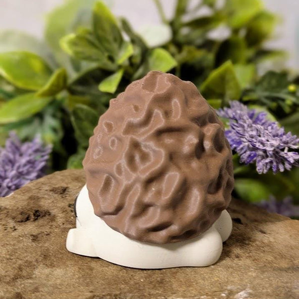 Side view of mushroom figurine with morel-style cap and white base, posed on stone with leafy floral backdrop.
