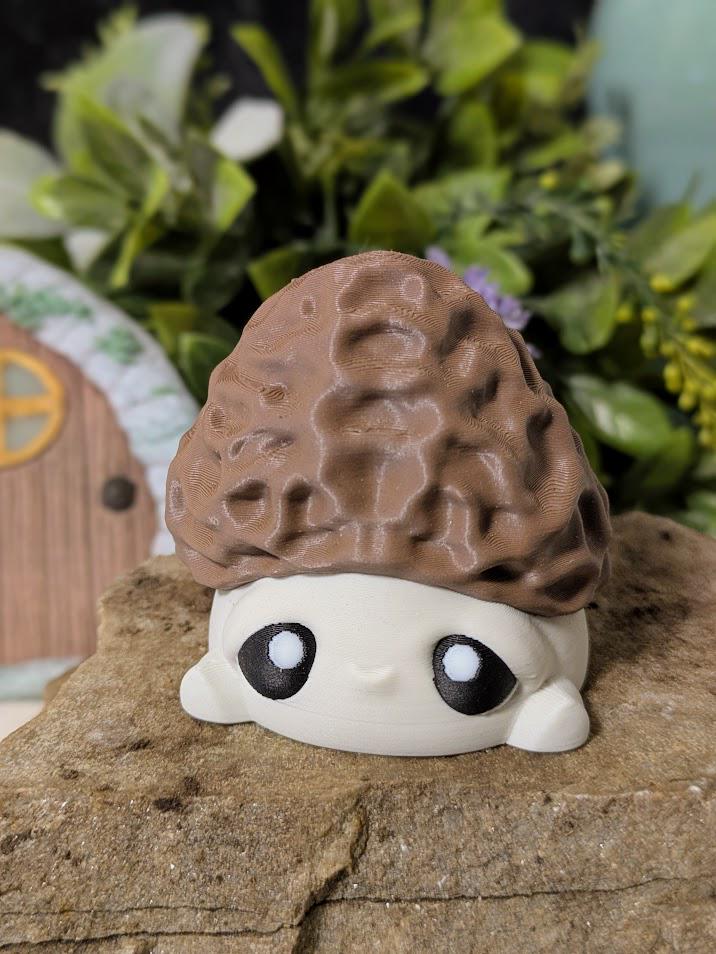 Front view of wide-eyed mushroom goblin with morel-style cap, sitting on stone with fairy door in background.