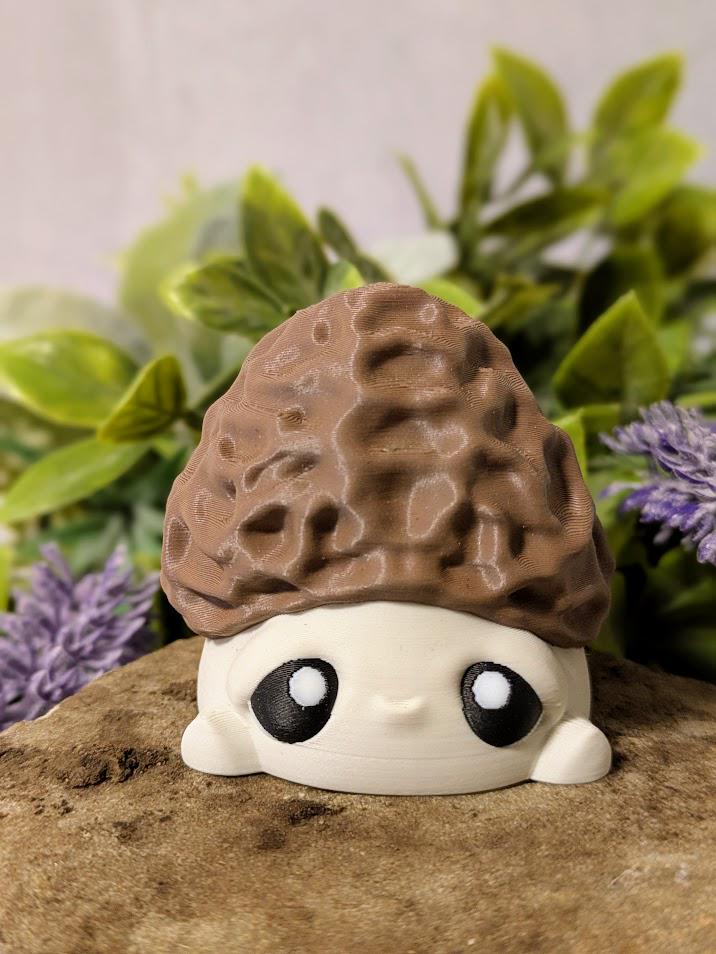 Straight-on view of mushroom goblin with morel-textured cap and round eyes, seated on a rock with florals behind.