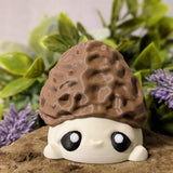 Straight-on view of mushroom goblin with morel-textured cap and round eyes, seated on a rock with florals behind.