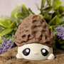 Straight-on view of mushroom goblin with morel-textured cap and round eyes, seated on a rock with florals behind.