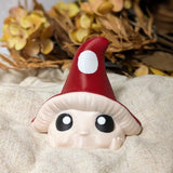 Red-capped mushroom goblin with big eyes, seated on a soft beige cloth with autumn leaves in the background.