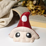 Front view of red-capped mushroom goblin with white spots and wide eyes, posed against dried flowers and an open book.