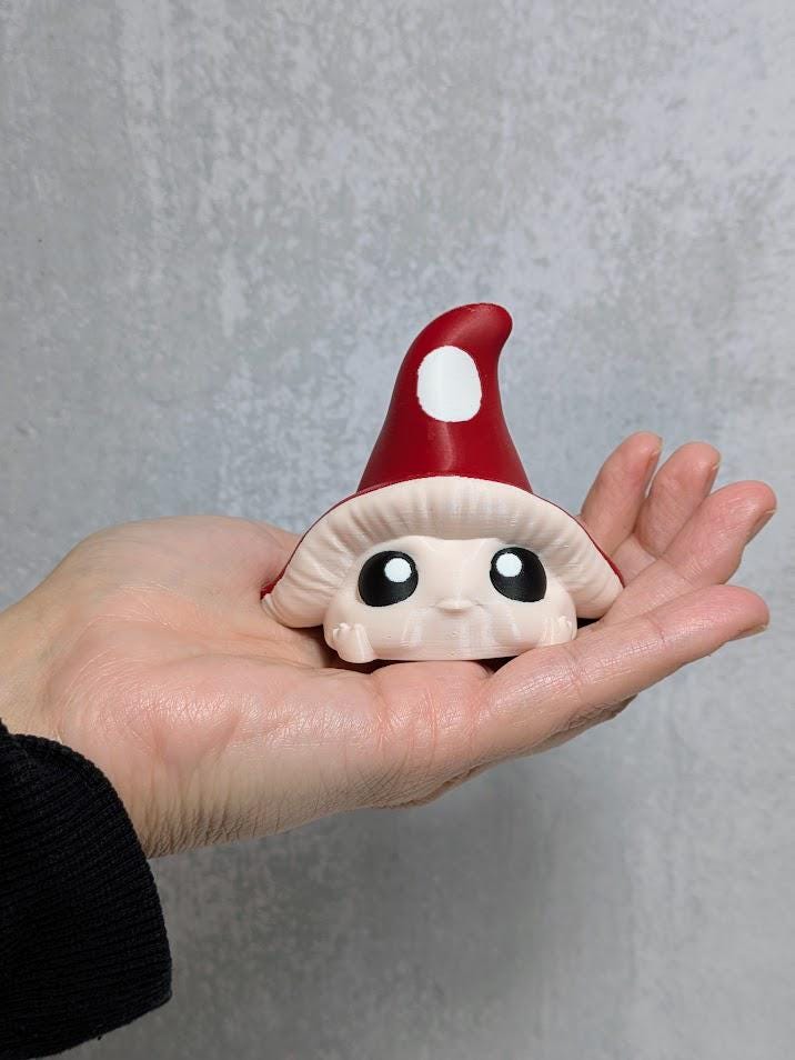 Red-capped mushroom goblin with wide eyes resting in an open hand for size reference, against gray background.