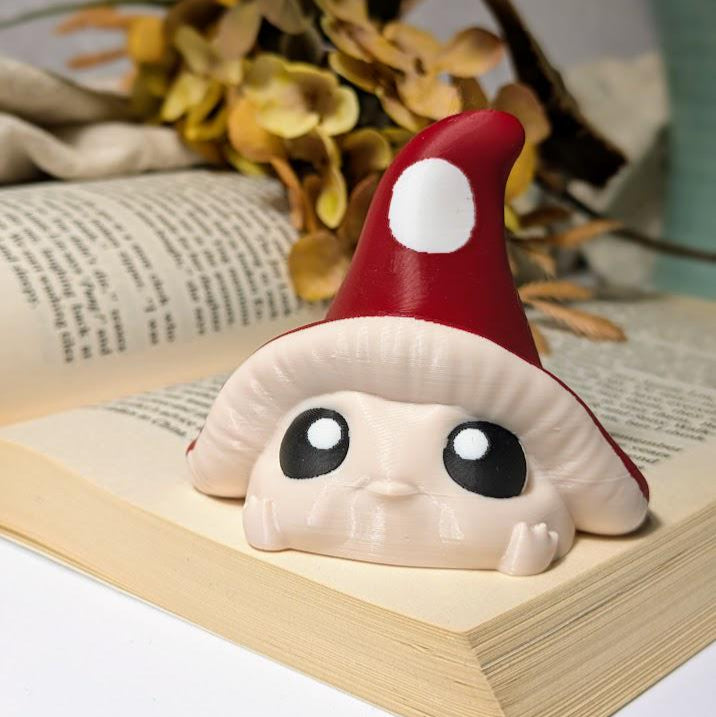 Mushroom goblin with red cap and white spots resting on open book with fall leaves in background.