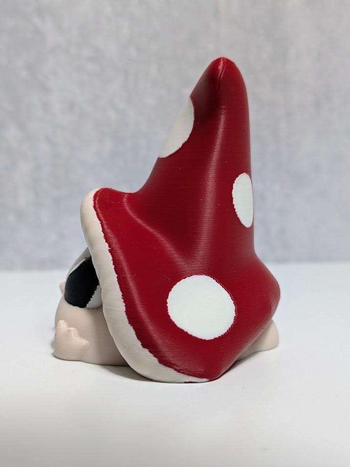 Side view of red-capped mushroom goblin with large white spots, partially hiding behind its oversized hat.