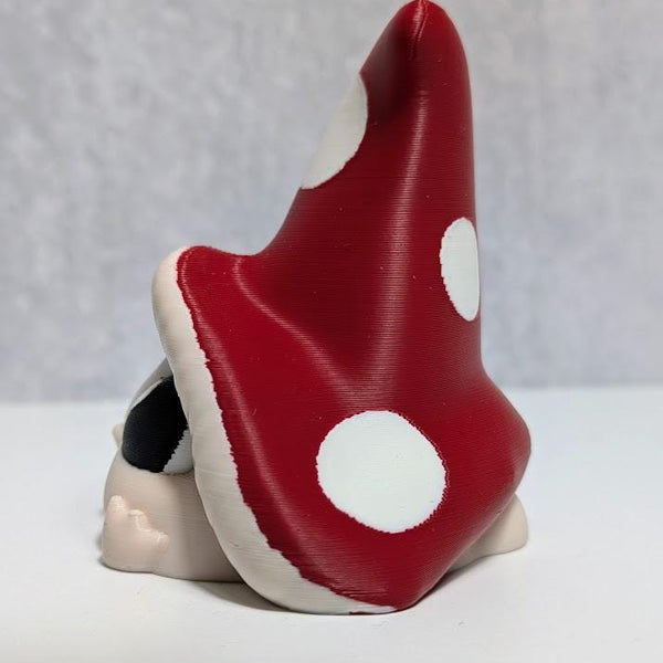 Side view of red-capped mushroom goblin with large white spots, partially hiding behind its oversized hat.